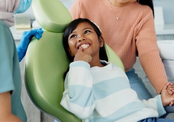 Girl sitting in a dentist's chair with her mouth open, while a woman wearing gloves stands beside her
