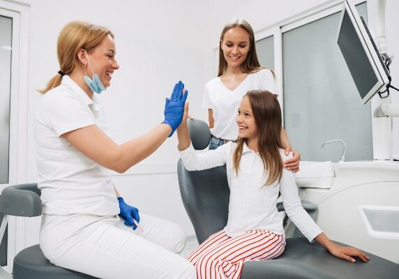 Woman sitting in a dentist's chair with her daughter, both of them The woman is wearing a mask and gloves