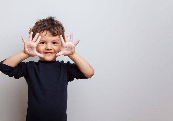 Boy standing in front of a white wall with his hands up in the air, wearing a t-shirt