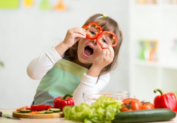 Little girl sitting at a table with a variety of vegetables, including capsicum, leafy vegetables, a chopping board, a knife