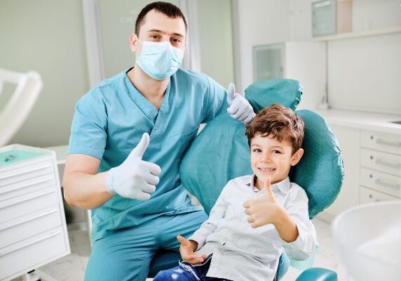 Boy sitting in a dentist's chair with his father giving a thumbs up