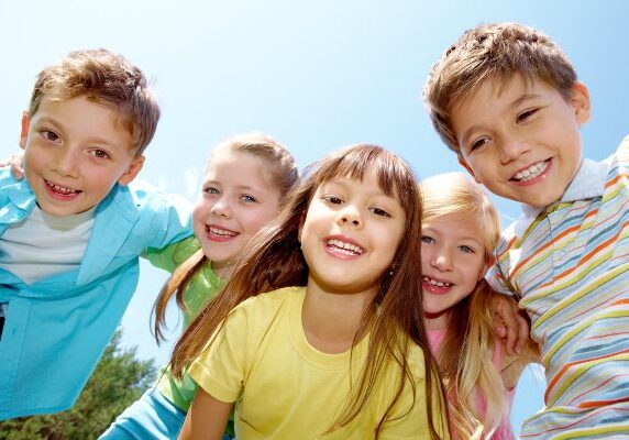 Group of children standing next to each other in a park, and surrounded by trees and a clear blue sky